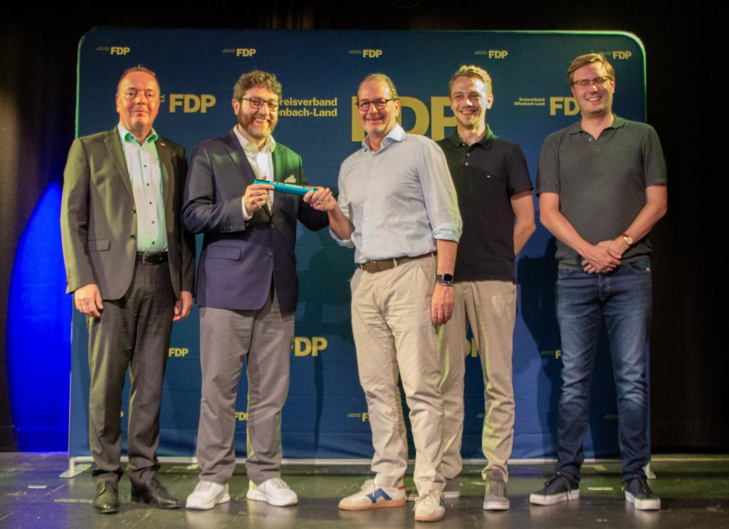 Fünf Mitglieder der FDP bei einer Staffelstabübergabe vor einer FDP-Fotowand des Kreisverbands Offenbach-Land.